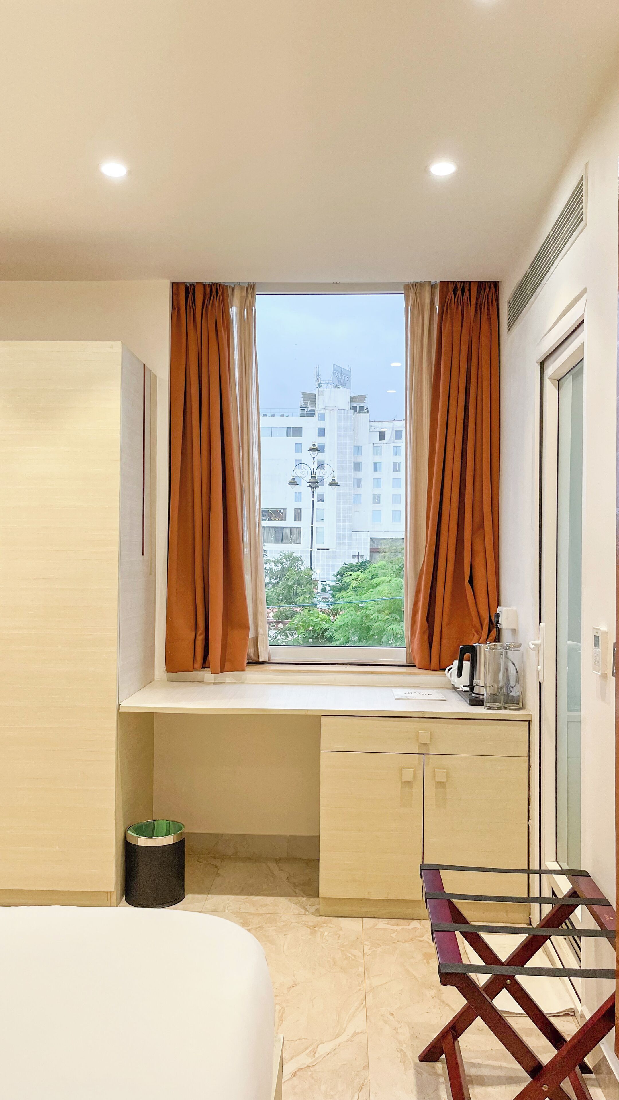 Habitación de hotel con cama, escritorio con computadora, ventana con cortinas, un cesto de basura y vista a edificios.