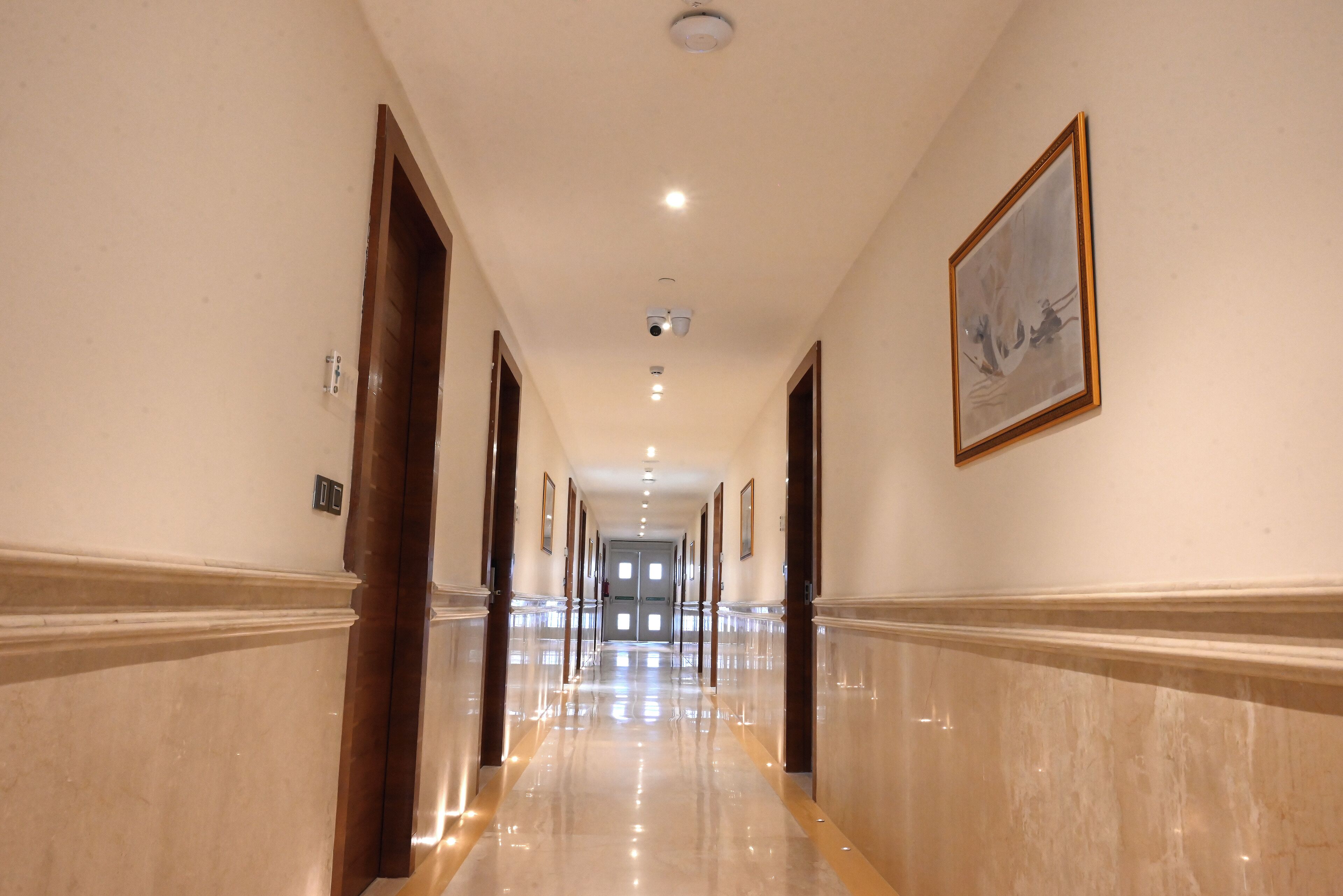 A long, well-lit hallway with marble floors and wooden doors, leading to a double door at the end.