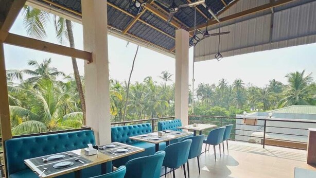 A restaurant with blue upholstered booths and chairs, a tiled floor, and a view of palm trees. - Singing Sands Beach Resort (Alibag)