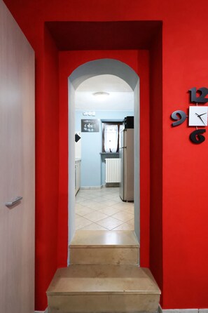 A hallway with a red wall, a clock, and a kitchen area with a refrigerator and a microwave. - Vacation Home King Cit (Diano d'Alba)