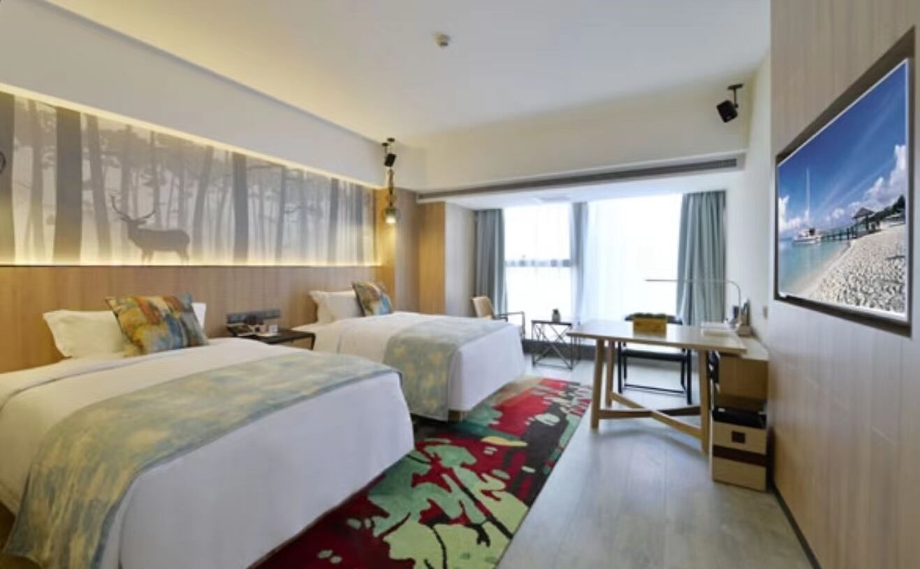 Ett hotellrum med två sängar, ett skrivbord, en stol, ett stort fönster och en tavla som föreställer en strandvy.