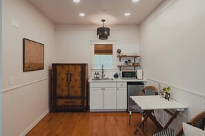 A compact kitchen with white cabinetry, a wooden cabinet, a microwave, and a dining table with chairs. - Swank Suites | Step Free King - Pet Friendly (Ashland)