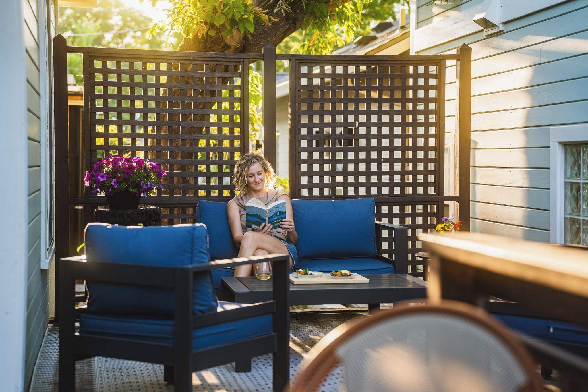 A person sitting on a blue sofa in a covered outdoor area with a wooden lattice partition and a table with a drink.