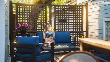 A person sitting on a blue sofa in a covered outdoor area with a wooden lattice partition and a table with a drink.