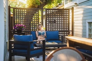 A person sitting on a blue sofa in a covered outdoor area with a wooden lattice partition and a table with a drink. - Swank Suites | Step Free King - Pet Friendly (Ashland)