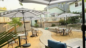 Un patio al aire libre con varias mesas y sillas, con sombrillas a rayas, rodeado por una terraza de madera y un camino de ladrillo.