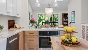 A modern kitchen with white cabinets, a built-in dishwasher, a large window, and a wooden island with fruit.