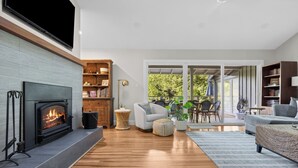 A modern living room with a fireplace, wooden flooring, and a large window overlooking a garden.
