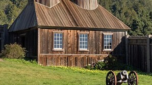 A rustic wooden church with a bell tower and a small steeple, surrounded by a wooden fence and a cart in the foreground.