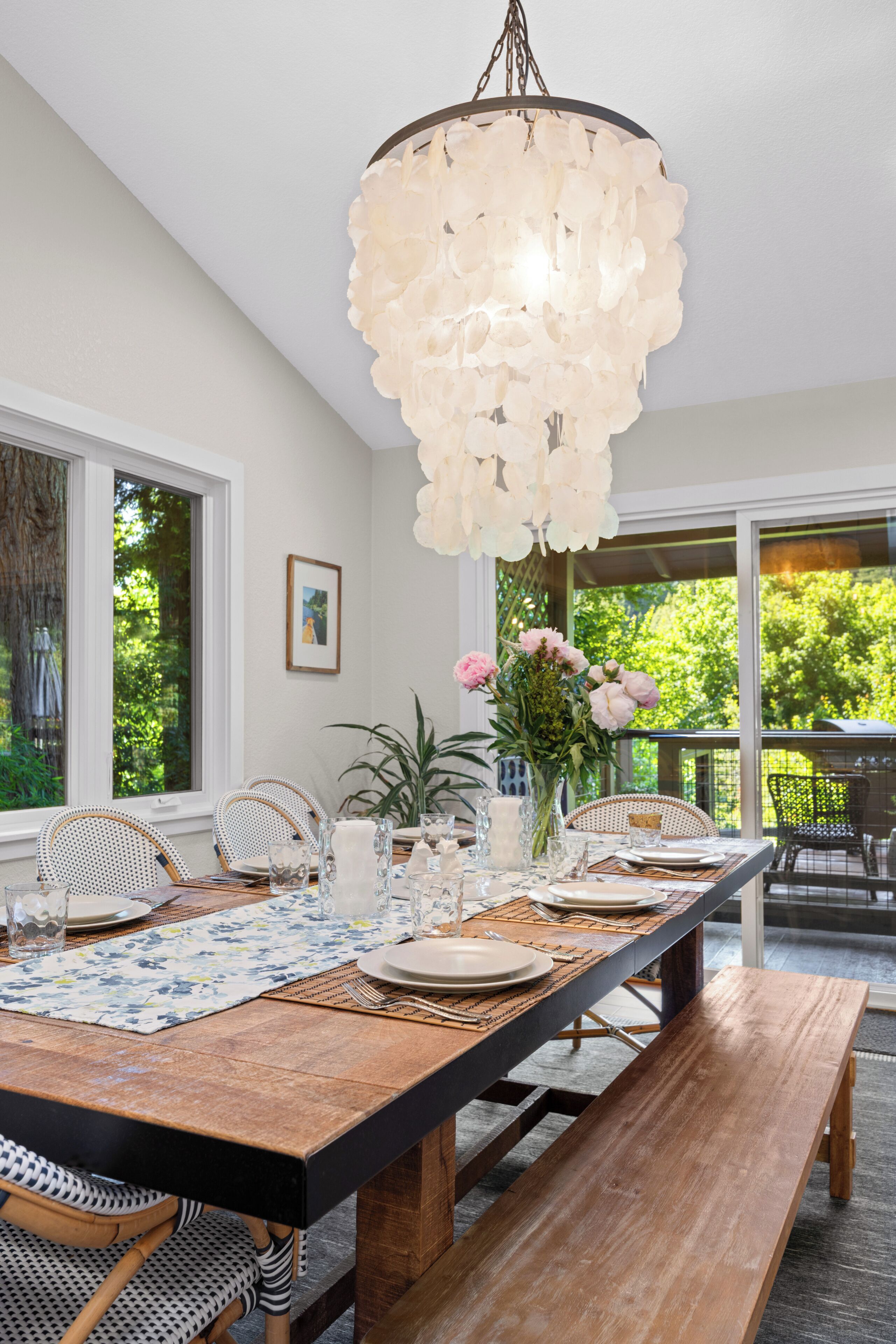 Un comedor con una larga mesa de madera, sillas, una lámpara colgante con flores y vistas a zonas verdes al exterior.