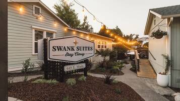 A sign for Swank Suites with a street address, surrounded by a landscaped area with mulch, small plants, and a wooden deck.