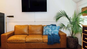 Un canapé en cuir marron agrémenté d’un plaid bleu et d’un coussin à motifs, une grande plante en pot, un téléviseur à écran plat fixé au mur et une lampe sur pied.