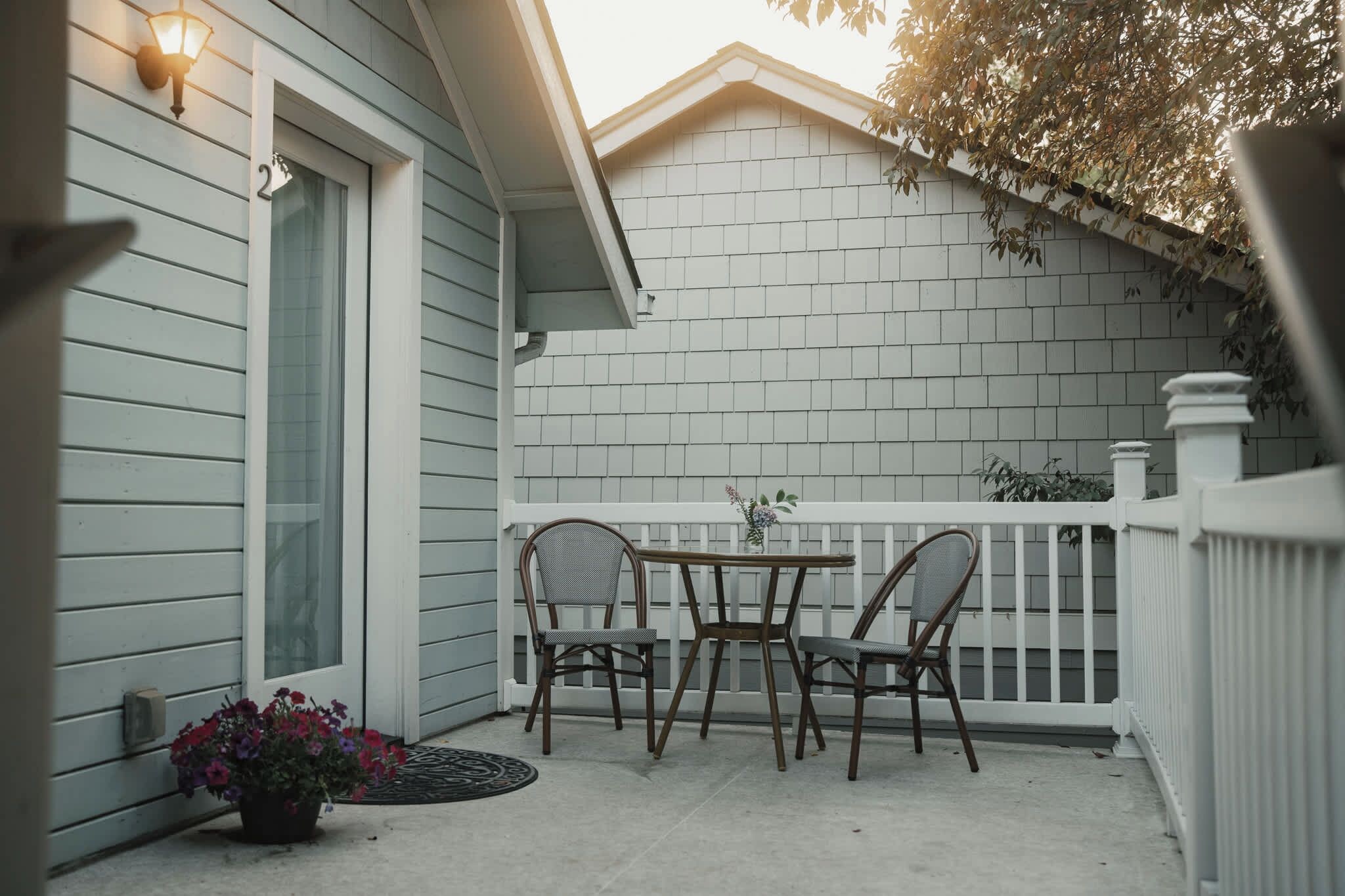 A covered patio with a table and two chairs, a potted plant with flowers, and a white fence.