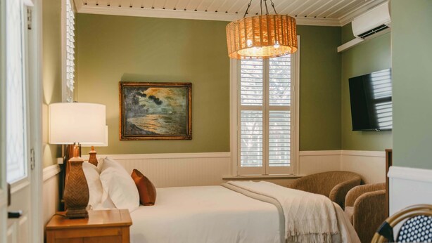 A hotel room with a bed, a chair, a table, a lamp, a painting, and a TV on the wall. - Swank Suites + Private Porch Patio (Ashland)