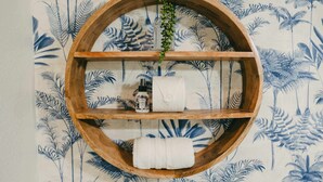 A wooden shelf with a glass container, a bottle, and a towel against a wallpaper with a blue and white tropical leaf pattern. - Swank Suites + Private Porch Patio (Ashland)