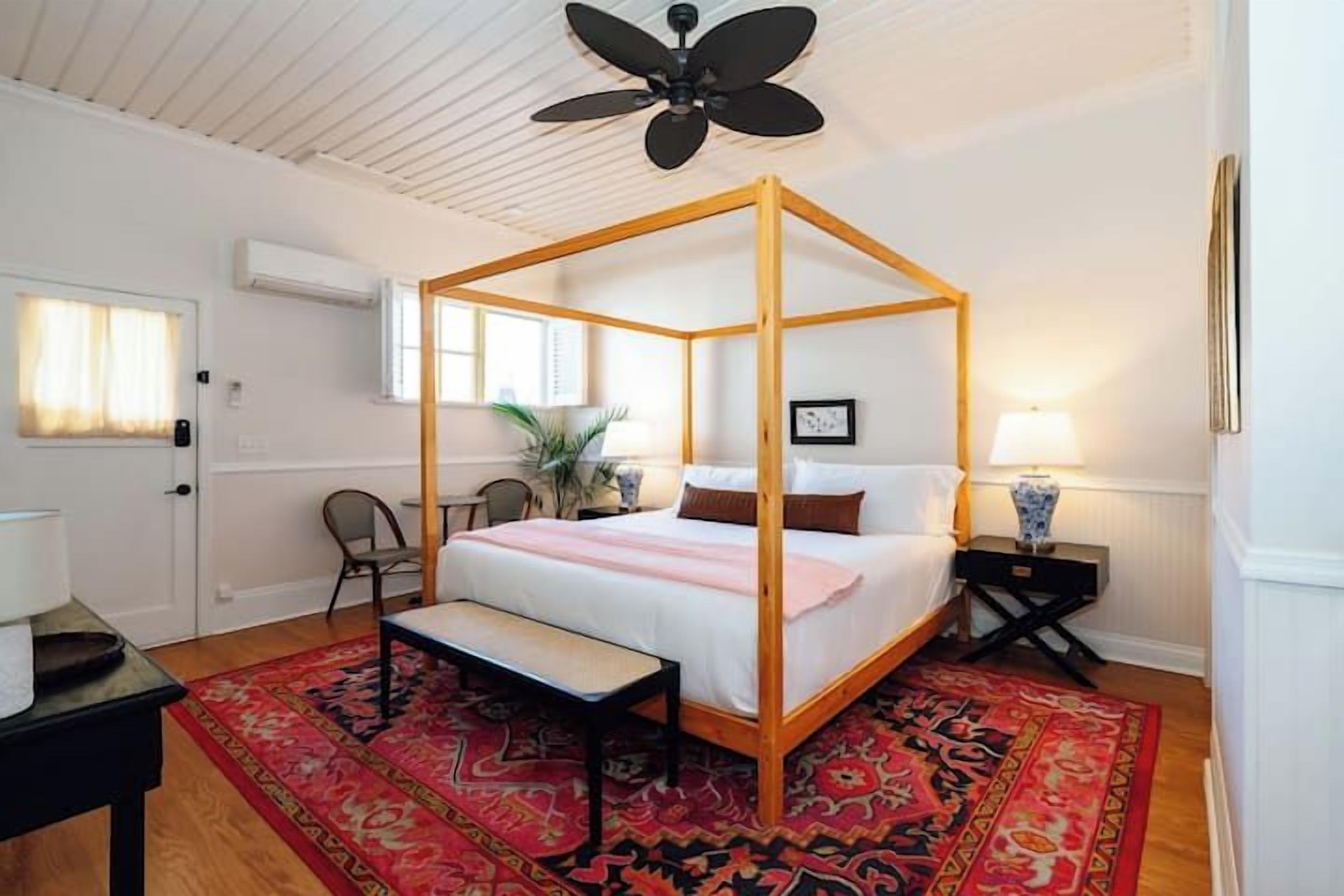 Een hemelbed met houten frame, een nachtkastje met lamp en een rood tapijt met patroon.