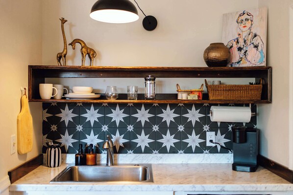 A kitchen with a marble countertop, a stainless steel sink, a coffee maker, and a decorative shelf with a giraffe figurine and a basket. - Swank Suites | King with Lux Tub (Ashland)