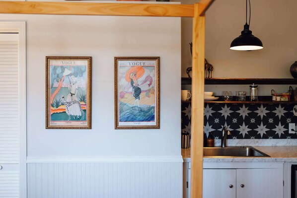 A kitchen with white cabinetry, a black backsplash with star patterns, and two framed vintage Vogue magazine covers on the wall.
