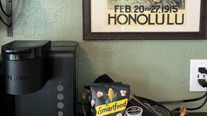A Keurig coffee maker, a basket with Smartfood and Stash snacks, and a framed poster of the Mid-Pacific Carnival in Honolulu. - Swank Boutique Suites | King Suite | DT Ashland (Ashland)