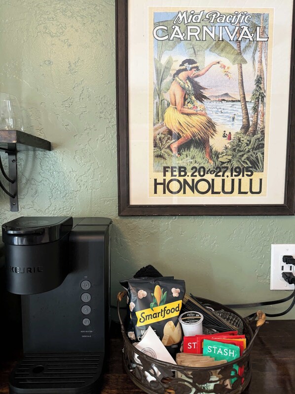 A Keurig coffee maker, a basket with Smartfood and Stash snacks, and a framed poster of the Mid-Pacific Carnival in Honolulu. - Swank Boutique Suites | King Suite | DT Ashland (Ashland)