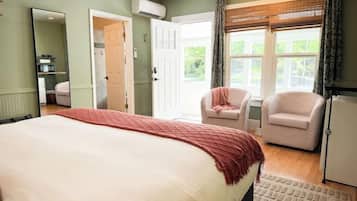 Un dormitorio con una cama grande, dos sillas, un ventanal con cortinas y una puerta que da a otra habitación.