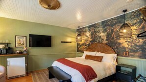 A bedroom with a bed, a desk, a refrigerator, a bench, and a mural on the wall. - Swank Boutique Suites | King Suite | DT Ashland (Ashland)