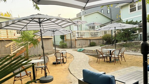 An outdoor patio area with multiple tables and chairs, shaded by striped umbrellas, and surrounded by a wooden deck and a brick pathway. - Swank Boutique Suites | King Suite | DT Ashland (Ashland)