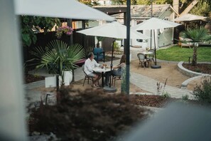 Un patio exterior con mesas y sillas bajo sombrillas, rodeado de plantas en macetas y una valla de madera.