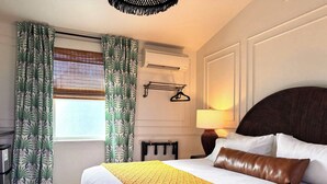 A hotel room with a large bed, a bedside table with a lamp, a wall-mounted air conditioner, and a window with curtains. - Swank Suites | King with Lux Tub (Ashland)