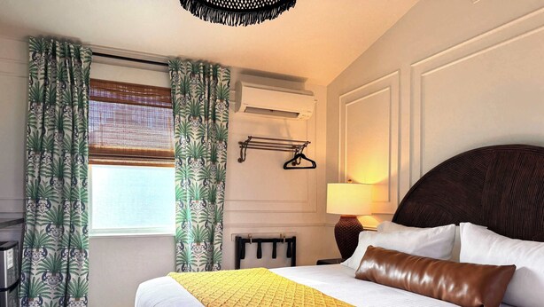 Habitación de hotel con una cama grande, una mesita de noche con lámpara, aire acondicionado de pared y una ventana con cortinas.