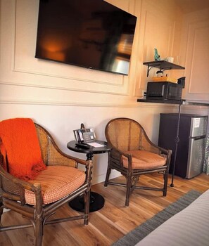 A hotel room with a flat-screen TV mounted on the wall, two wicker chairs with cushions, a small round table, and a refrigerator.