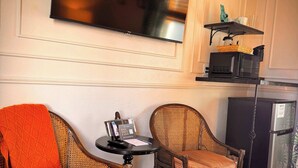 A hotel room with two wicker chairs, a small table with a book and a pen, a flat-screen TV mounted on the wall, a microwave, and a refrigerator. - Swank Suites | King with Lux Tub (Ashland)