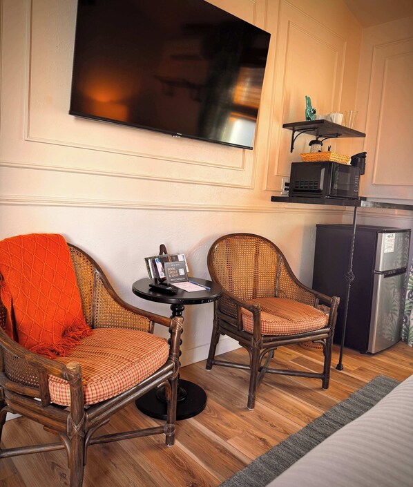 A hotel room with two wicker chairs, a small table with a book and a pen, a flat-screen TV mounted on the wall, a microwave, and a refrigerator.