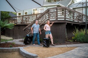 Una pareja con equipaje de pie frente a una casa con escalera de madera y un área cercada con plantas.