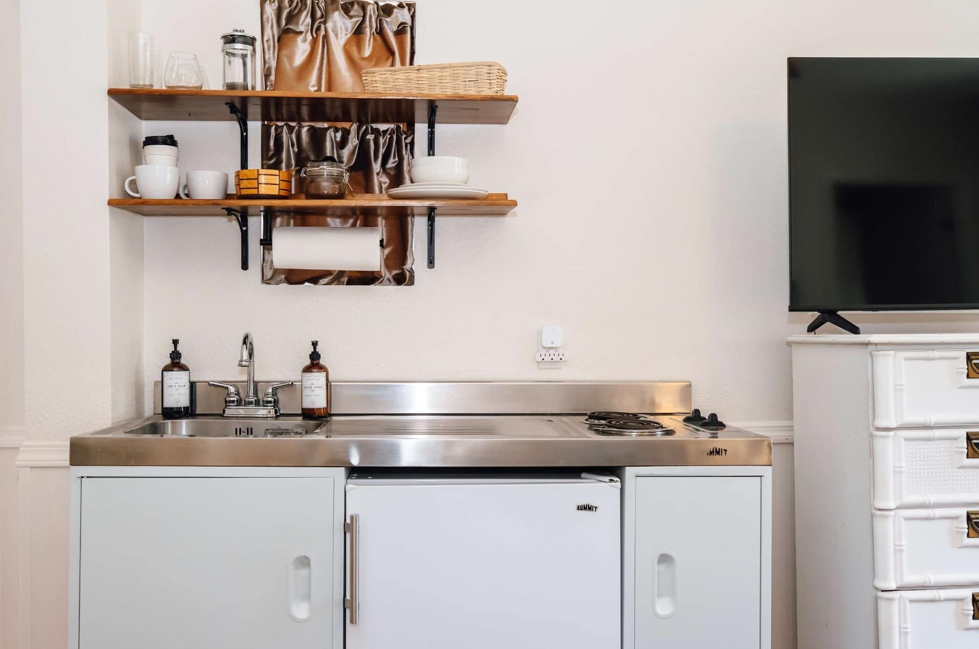 Una cocina con pileta de acero inoxidable, armarios blancos y un televisor de pantalla plana montado sobre los armarios.