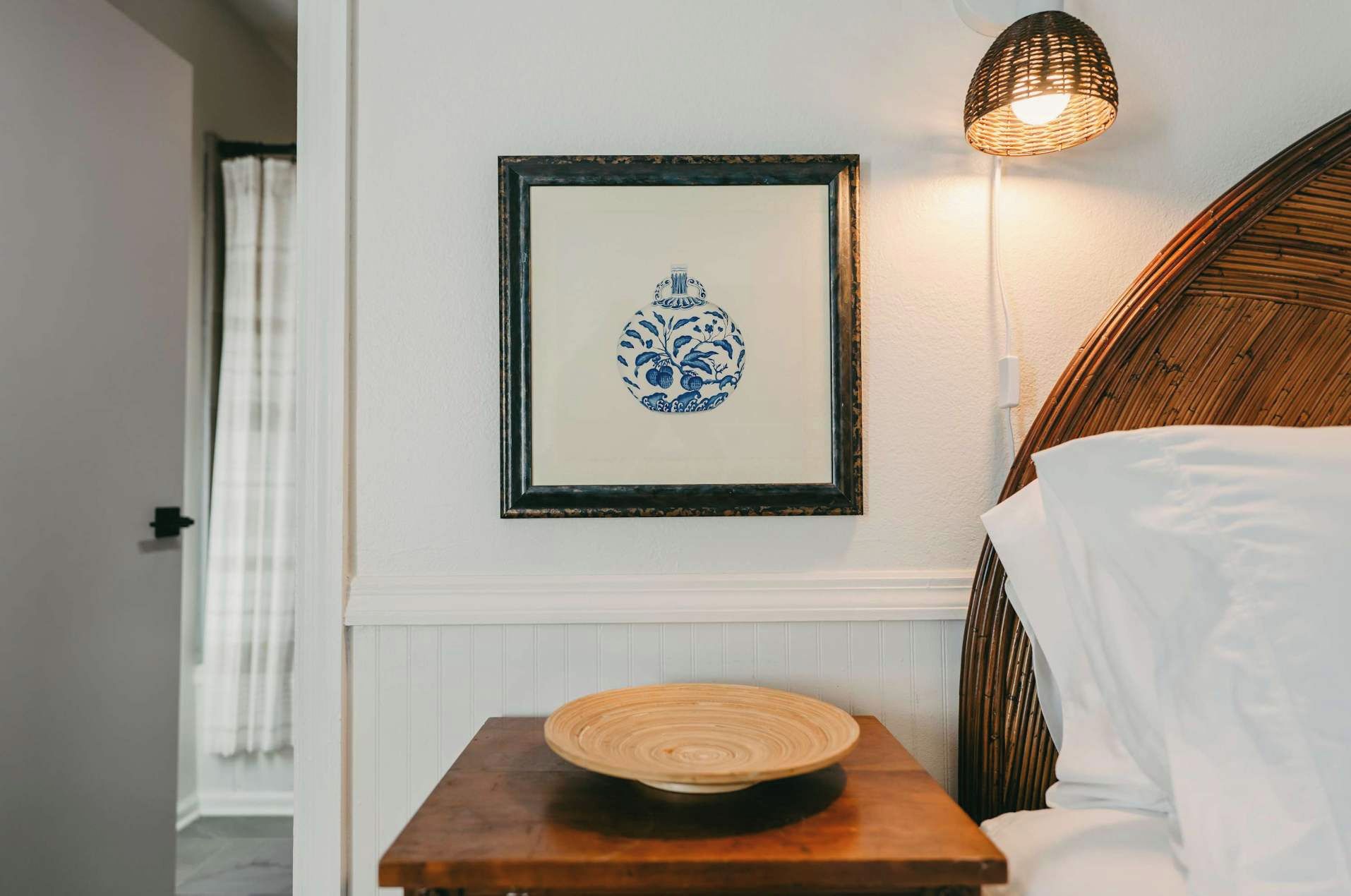 Una obra de arte enmarco con un diseño azul y blanco en una pared, una mesita de noche con un objeto redondo de madera, y una lámpara fijada a la pared.