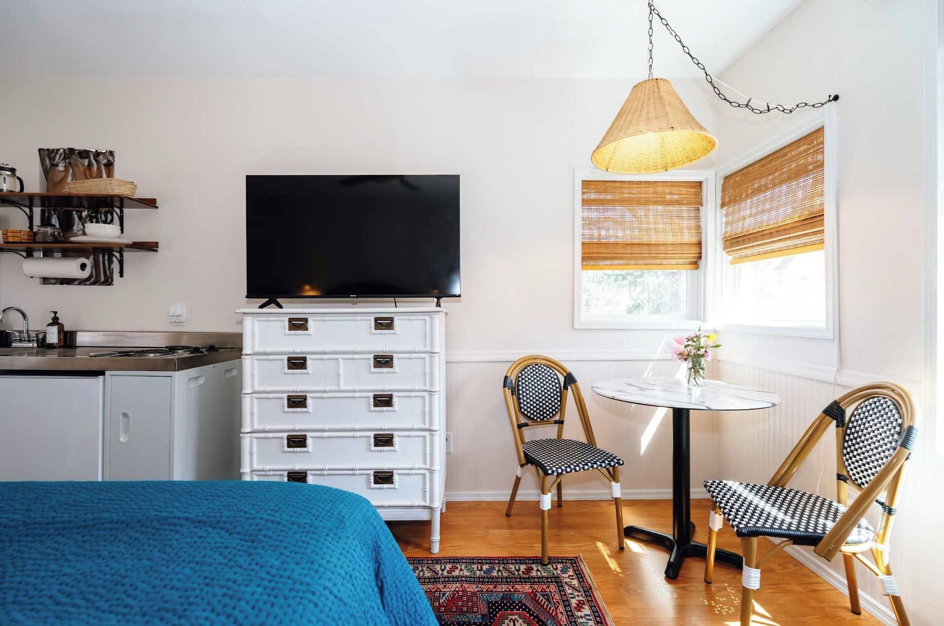 Habitación de hotel pequeña con cama, televisión, una mesa pequeña para comer, dos sillas, cómoda y ventana con persianas.