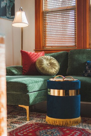 Ein Zimmer mit einem grünen Sofa, einem roten Kissen, einem gold- und blauem Hocker und einer Stehlampe.