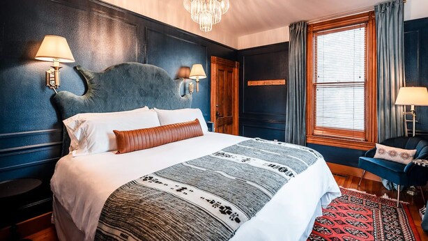 Ein Hotelzimmer mit einem großen Bett, zwei Wandlampen, einem Chandelier, einem Fenster mit Vorhängen und einem kleinen Tisch mit einem Stuhl.