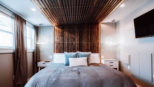 A modern bedroom with a large bed, wooden slatted ceiling, and a flat-screen TV on the wall.