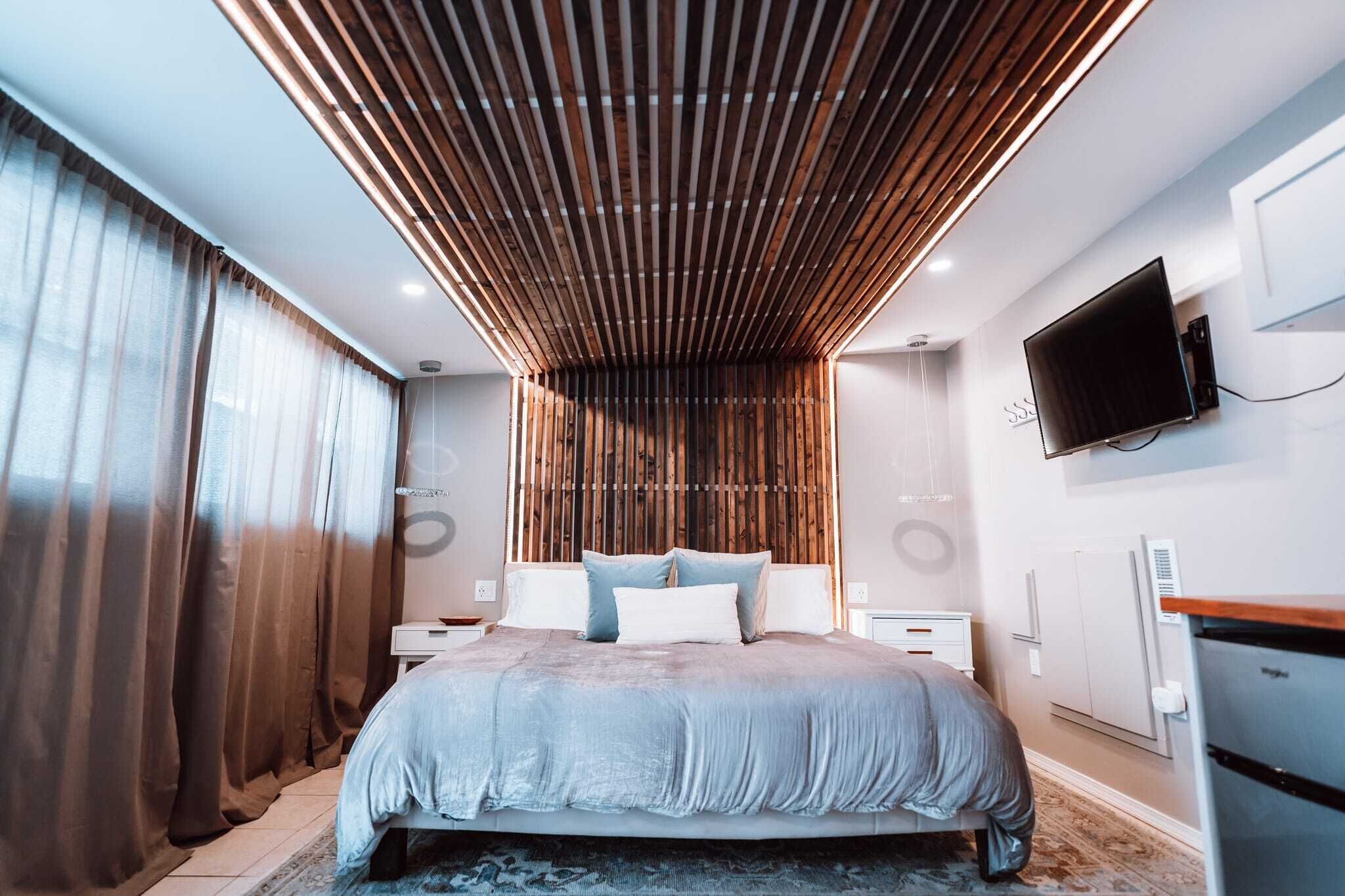 A modern bedroom with a large bed, wooden slatted ceiling, a flat-screen TV, and a built-in wardrobe.