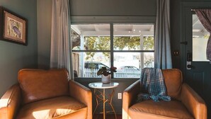 Een kamer met twee leren fauteuils, een kleine ronde tafel met een plant en een raam met uitzicht op een hek en bomen.