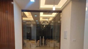 A modern reception area with a wooden reception desk, a potted plant, and a glass door leading to a conference room with tables and chairs. - Hotel Innvite (Agra)