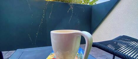 A white mug on a black table with a colorful book underneath, set against a backdrop of greenery and a clear sky.