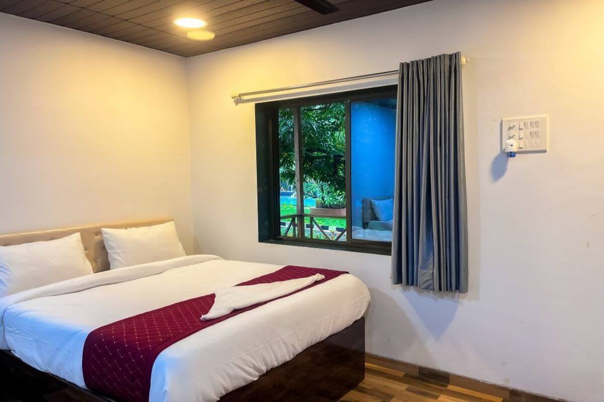 A hotel room with a bed, a window with curtains, and a view of greenery outside.