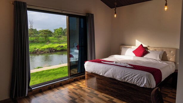 Ein Schlafzimmer mit einer großen Schiebeglastür, die auf einen Teich und viel Grün blickt. Der Raum hat einen Holzboden und ein Bett mit weißer Bettwäsche und einem roten Kopfkissen.