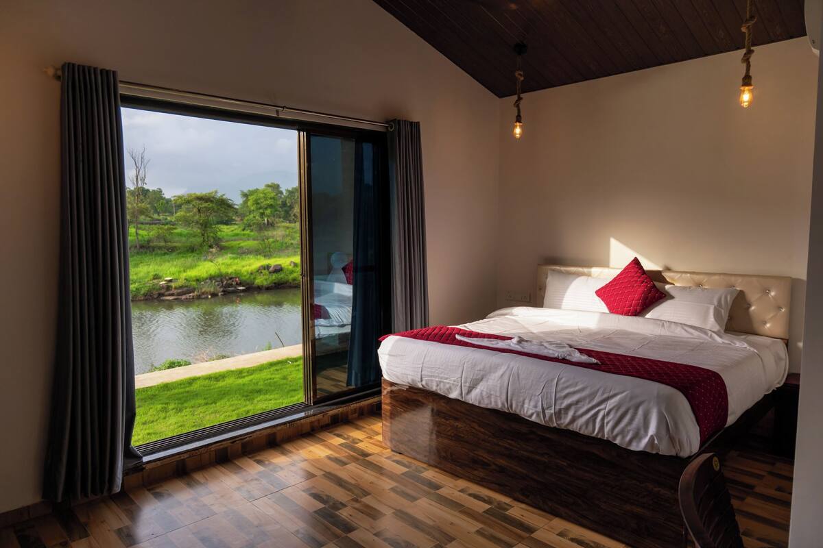 A bedroom with a large sliding glass door leading to a view of a pond and greenery. The room has a wooden floor and a bed with white bedding and a red pillow.
