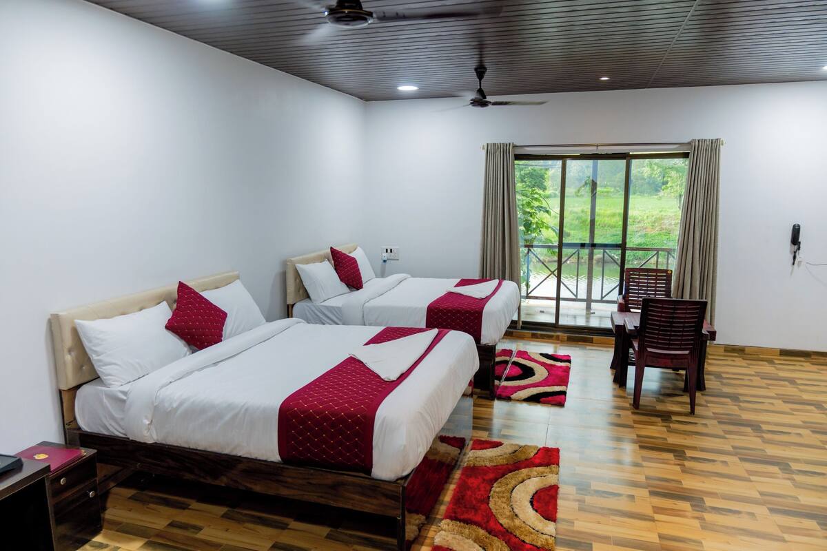 A hotel room with two beds, a dining table, and a view of greenery outside.