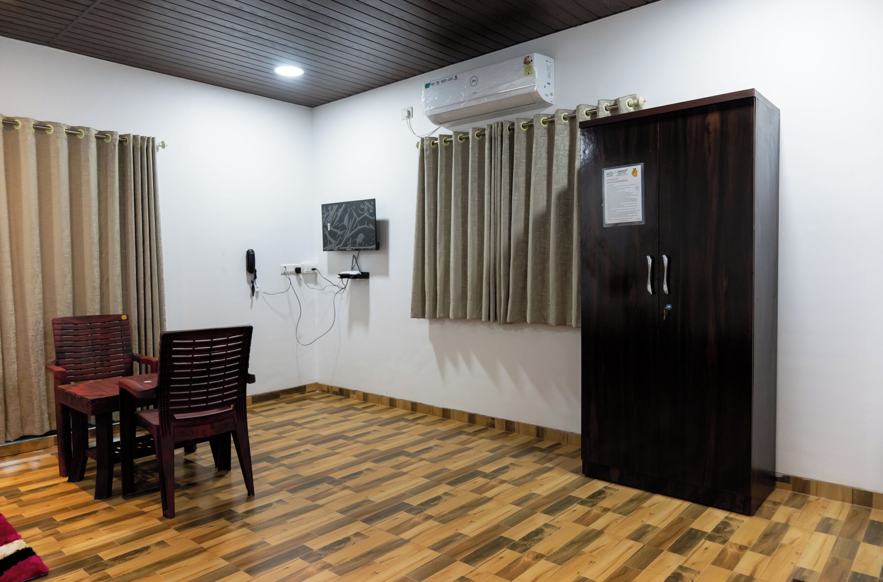 A room with a wooden parquet floor, a dark wooden wardrobe, a desk with two chairs, an air conditioning unit, and a wall-mounted television.