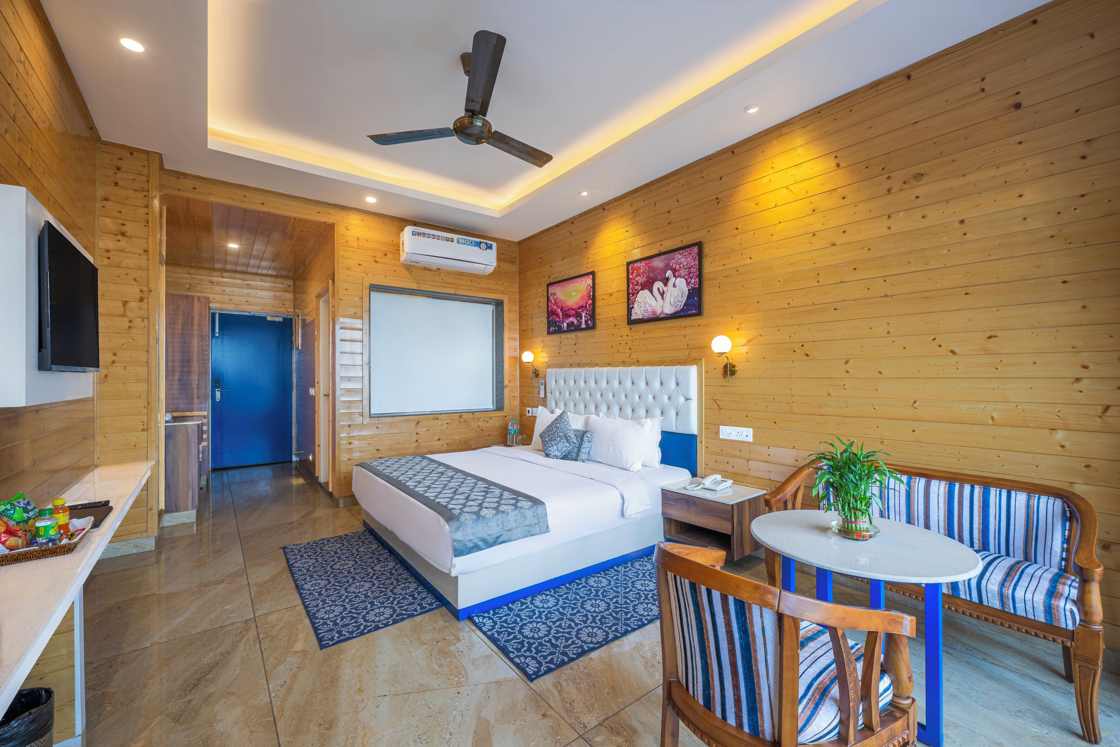 Une chambre d’hôtel moderne avec un intérieur en bois, un lit recouvert d’une courtepointe à motifs bleus et blancs, un ventilateur de plafond, une petite table ronde avec un pot de plante et un canapé à rayures.
